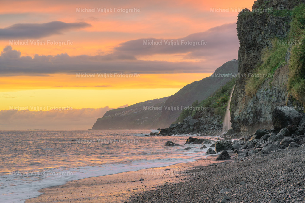 An der Küste in Ponta do Sol auf Madeira | Stellen Sie sich einen Ort vor, an dem die Zeit stillzustehen scheint, während die Sonne langsam am Horizont versinkt und den Himmel in leuchtenden Orange-, Rosa- und Goldtönen bemalt. Das ist Ponta do Sol auf Madeira, ein malerisches Küstendorf, das seinem Namen alle Ehre macht: "Spitze der Sonne".In diesem besonderen Moment zieht die Steilküste die Blicke auf sich. Rechts im Bild stürzt ein schlanker Wasserfall die steilen Felsen hinab und ergießt sich direkt ins Meer. Es ist ein Schauspiel der Elemente – das fließende Wasser trifft auf die endlose Weite des Ozeans, während die untergehende Sonne den Himmel in ein Farbenmeer verwandelt.Die Landschaft ist eine harmonische Mischung aus rauer Natur und beruhigender Schönheit. Die steilen Klippen, das sanfte Rauschen des Wassers und die Farben des Himmels schaffen eine Szene, die gleichzeitig Kraft und Frieden ausstrahlt.Ponta do Sol ist ein Ort, an dem man die Seele baumeln lassen kann. Dieser Sonnenuntergang lädt dazu ein, innezuhalten, durchzuatmen und den Moment in vollen Zügen zu genießen – ein perfekter Augenblick, um die Schönheit der Natur in ihrer reinsten Form zu erleben. - Realisiert mit Pictrs.com