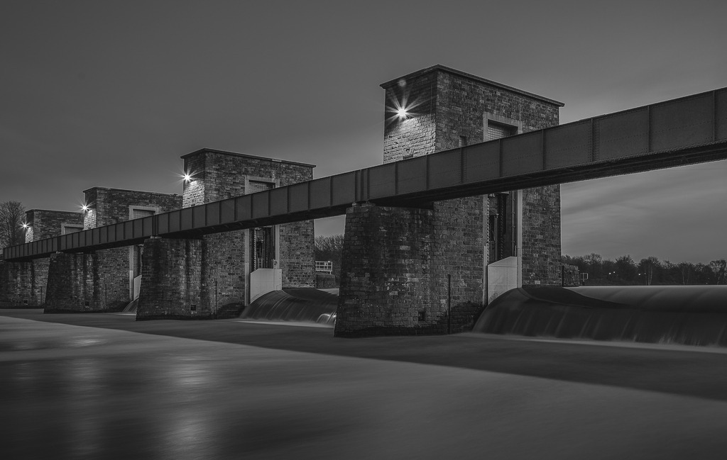 Industriewehr in Schwarzweiß | Diese minimalistische Schwarzweißaufnahme zeigt eine industrielle Wehranlage als grafisch klare Fine-Art-Komposition. Geometrische Linien, wiederkehrende architektonische Elemente und das weich fließende Wasser der Langzeitbelichtung erzeugen eine stille, zugleich kraftvolle Bildwirkung.Stahl, Stein und Wasser verbinden sich zu einer zeitlosen urbanen Szenerie, in der technische Strenge auf fließende Bewegung trifft. Feine Lichtreflexe und ruhige Tonwerte unterstreichen den spannungsvollen Kontrast zwischen Konstruktion und Dynamik und verleihen dem Motiv seine markante, fast skulpturale Präsenz. - Realisiert mit Pictrs.com