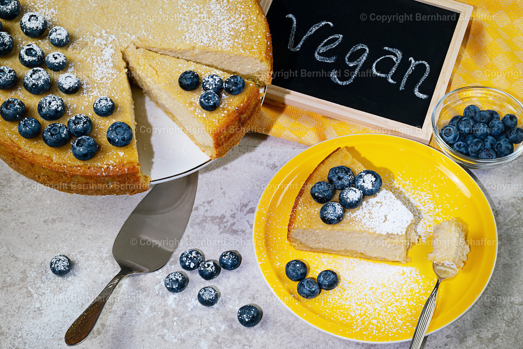 Käsekuchen vegan | Ein veganer Käsekuchen mit Blaubeeren, ein Stück Kuchen auf einem Teller, ein Sachale mit Blaubeeren und eine Kreidetafel mit der Aufschrift „vegan“ von oben fotografiert. - Realisiert mit Pictrs.com