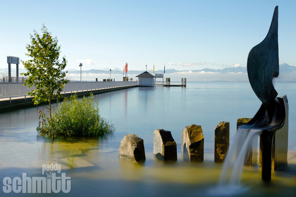 Brunnen am Landesteg bei Immenstaad am Bodensee | Ein idyllischer Brunnen am Landesteg bei Immenstaad verbindet maritime Atmosphäre mit dem besonderen Charme des Bodenseeufers. Im Hintergrund eröffnet sich der Blick auf den See und lässt die ruhige, entspannte Stimmung dieses Ortes spürbar werden. - Realisiert mit Pictrs.com