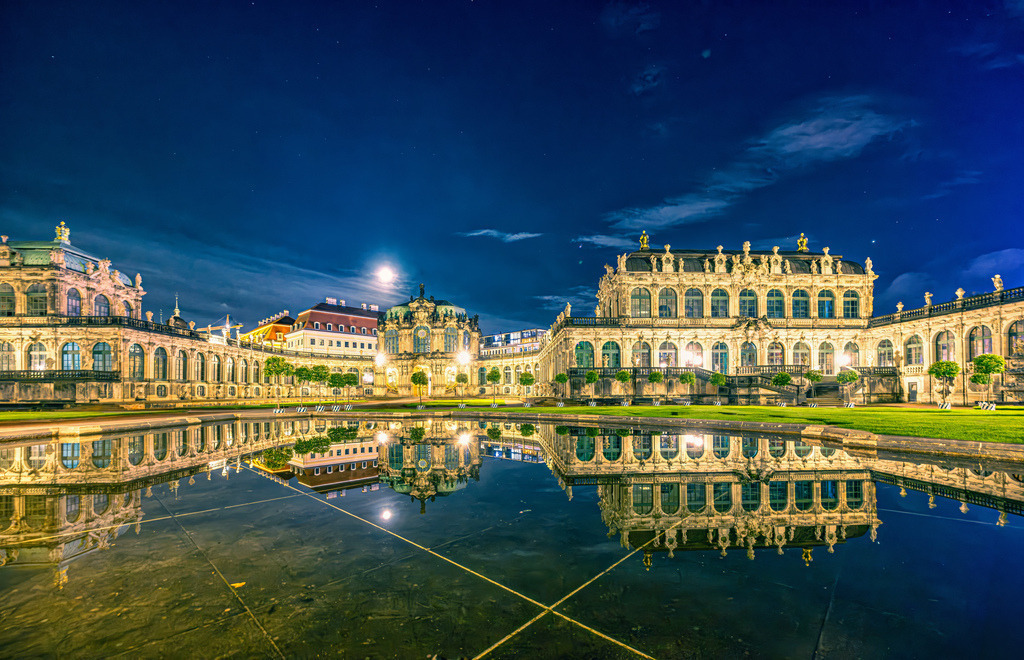 Nachtaufnahme des berühmten Innenhof des Dresdner Zwingers,barocker Gebäudekomplexes spiegelt sich im Brunnenbecken | Der markierte Bereich zeigt den berühmten Innenhof des Dresdner Zwingers, ein Meisterwerk der barocken Architektur im Herzen von Dresden. Das Foto fängt die beeindruckende abendliche Beleuchtung des historischen Gebäudekomplexes ein. Zu sehen sind die eleganten Pavillons und Galerien, die sich makellos in der großen, ruhigen Wasserfläche eines der Brunnenbecken spiegeln. Der Dresdner Zwinger wurde im frühen 18. Jahrhundert unter Kurfürst Friedrich August I. (bekannt als August der Starke) errichtet und beherbergt heute weltberühmte Museen wie die Gemäldegalerie Alte Meister und die Porzellansammlung. - Realisiert mit Pictrs.com