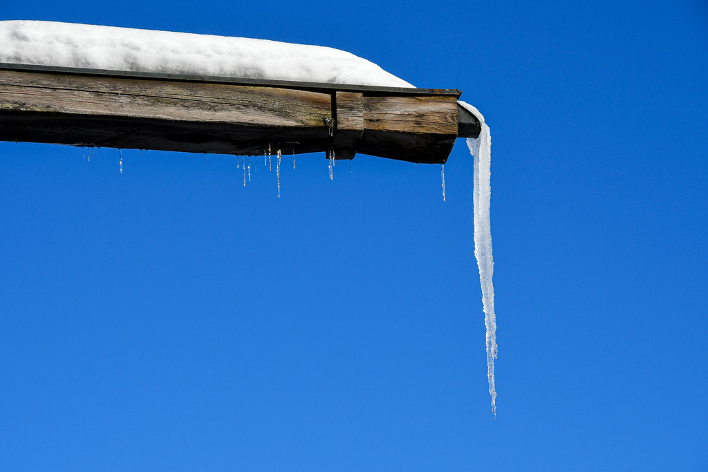 Kristallene Winterkunst: Eiszapfen an verschneitem Holzdach | Ein lichter und eiskalter Wintertag in Garmisch-Partenkirchen prägt dieses grafische Naturmotiv: Ein eindrucksvoller, langer Eiszapfen ragt steil von einer dicken Schneeschicht am Dachvorsprung eines alten Holzgebäudes herab. Vor dem makellosen, strahlend blauen Himmel setzt die filigrane Eisskulptur zusammen mit den kleineren Nachbarzapfen einen faszinierenden Akzent. Das Motiv vereint alpine Winterromantik, frostige Klarheit und die ruhige Poesie eines sonnigen, kalten Tages in den Bergen. - Realisiert mit Pictrs.com