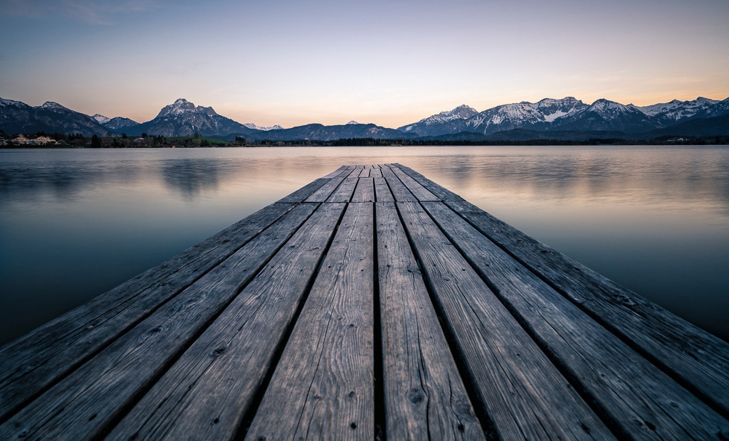 Perspektive der Stille – Steg am Hopfensee | Ein stiller Abend am Hopfensee im Allgäu – das Wasser liegt glatt wie Glas, die Berge spiegeln sich im sanften Licht der Dämmerung. Der hölzerne Steg zieht den Blick in die Ferne und lädt dazu ein, innezuhalten. Dieses Bild strahlt Ruhe und Weite aus – ein Moment der völligen Entschleunigung inmitten der Natur. - Realisiert mit Pictrs.com