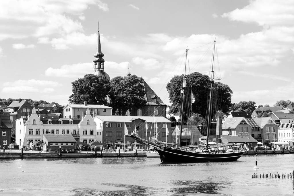 Traditionssegler im Hafen von Kappeln | Erleben Sie die historische Pracht von Kappeln mit dieser faszinierenden Schwarz-Weiß-Aufnahme. Das Bild zeigt einen majestätischen Traditionssegler, der durch den Hafen gleitet. Im Hintergrund sind die charmanten Gebäude der Stadt zu sehen, darunter der markante Kirchturm, der sich stolz gegen den Himmel erhebt und das historische Erbe der Stadt widerspiegelt.Dieses Kunstwerk, erhältlich auf Leinwand, Alu-Dibond, Acrylglas, Fineart und als Akustikbild, bringt die maritime Tradition und die architektonische Schönheit von Kappeln direkt in Ihr Zuhause. - Realisiert mit Pictrs.com