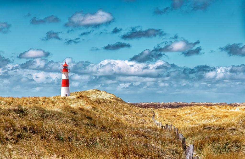 Leuchtturm List-Ost-Sylt-Ellenbogen-umrahmt von Strandhafer 2 | Der Leuchtturm List-Ost befindet sich auf der Halbinsel Ellenbogen im Norden von Sylt.Er wurde 1857 erbaut und ging am 1. Januar 1858 in Betrieb.Der Turm ist rot-weiß geringelt und seit 1977 ferngesteuert.Die Umgebung ist bekannt für ihre weite Dünenlandschaft und ist ein beliebtes Ziel für Wanderungen. - Realisiert mit Pictrs.com