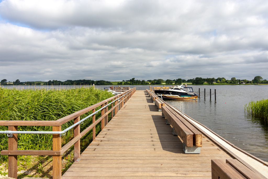 Wandbild: Steg in Sieseby an der Schlei | Dieses Wandbild im Querformat zeigt den neuen Steg in Sieseby im Sommer 2024. Links neben dem Steg ist Schilf zu sehen. In der Ferne kann man einige Boote am Steg erkennen. Der Himmel ist überwiegend bewölkt. - Realisiert mit Pictrs.com