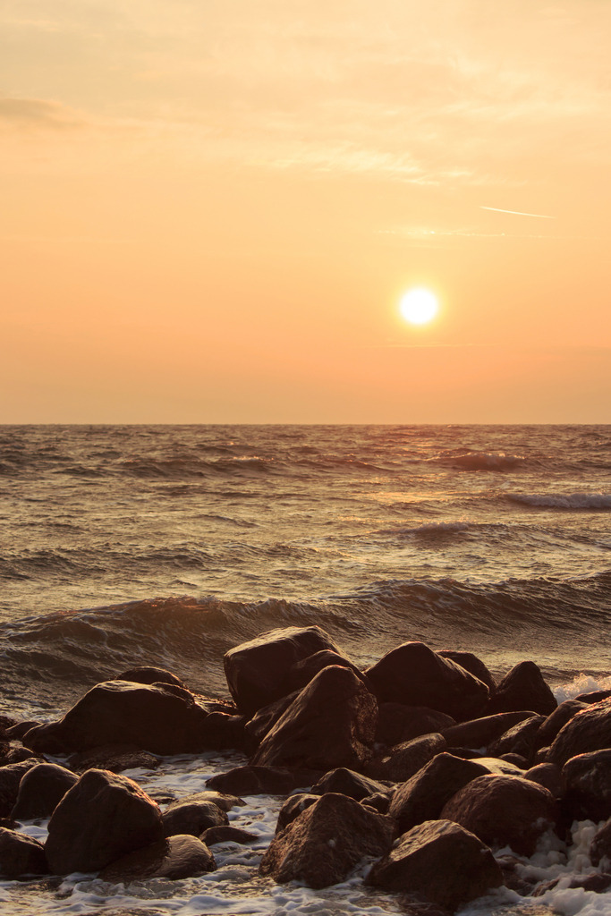 Wandbild: Sonnenaufgang an der Ostsee | Dieses Wandbild im Hochformat zeigt einen intensiven Sonnenaufgang am Strand von Damp. Der Himmel leuchtet in kräftigem Orangerot, während die Sonne über dem Horizont aufsteigt und das Meer sanft erhellt. Im Vordergrund liegen dunkle Steine der Steinmole, an denen sich leichte Wellen brechen. Die Szene vermittelt Ruhe, Weite und die kraftvolle Schönheit eines neuen Tages – ideal für Wohnräume, Ferienunterkünfte oder Praxisräume mit maritimer Atmosphäre. Erhältlich als Leinwand, Acrylglas, Alu-Dibond, hochwertiger FineArt-Druck oder als schallabsorbierendes Akustikbild. - Realisiert mit Pictrs.com