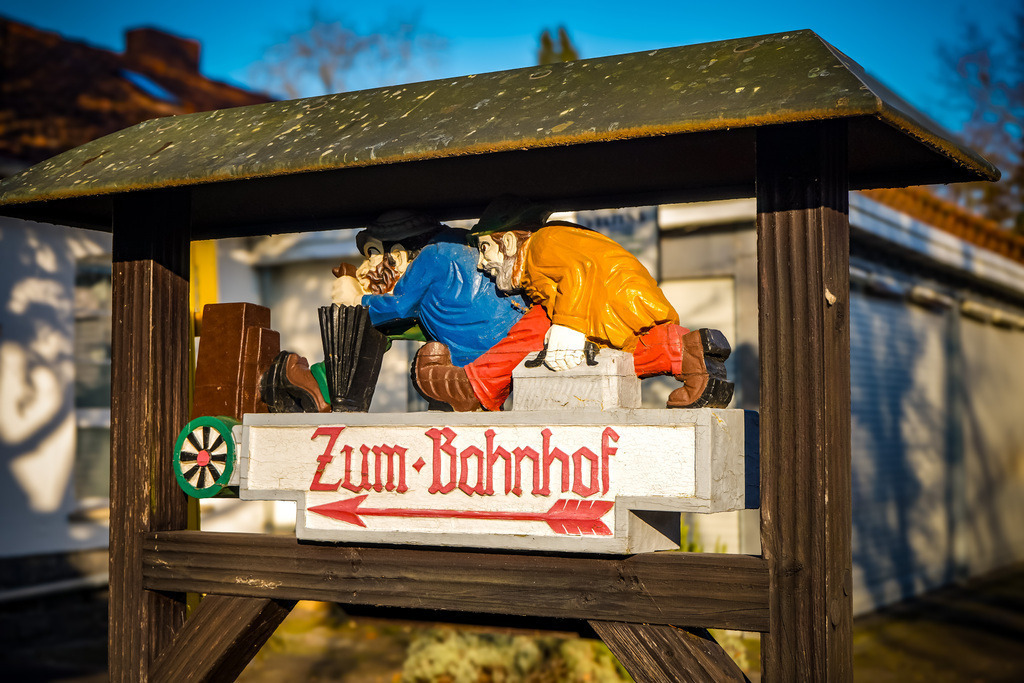 Zum Bahnhof - Bad Salzhausen | Ein verspieltes Hinweisschild lockert die Atmosphäre auf und sorgt sicher für den ein oder anderen schmunzelnden Seitenblick.'Der Kurpark in Bad Salzhausen ist vor allem für die mineralischen Quellen, das Gradierwerk und die Mühle der ehemaligen Salzsieder bekannt. Doch auch Abseits dieser Wasserattraktionen finden sich kleine und große Kunstwerke, die auch Stammgäste noch überraschen können. Man muss lediglich die angelegten Pfade mit wachem Auge begehen und als Lohn eröffnet sich eine neue Perspektive auf die grüne Kuranlage. - Realisiert mit Pictrs.com