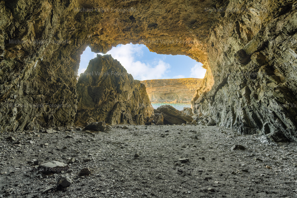 Höhlen von Ajuy auf Fuerteventura | Der Blick aus der Höhle von Ajuy führt aus dem tiefen Schatten hinaus auf die hellen, warmen Felswände der Küste. Die unregelmäßigen Konturen des Lavagesteins rahmen den Ausgang wie ein natürlicher Rahmen, während draußen das Sonnenlicht die Schichten des Gesteins klar modelliert. Die Helligkeit wirkt fast grell im Kontrast zur dunklen Höhle, und die Struktur des Felses tritt deutlich hervor. Ein ruhiger Moment, der die Stille im Inneren mit der klaren, sonnengetränkten Landschaft vor der Höhle verbindet. - Realisiert mit Pictrs.com