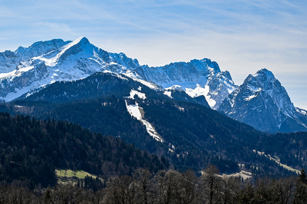 Wo die Erde den Himmel berührt – Garmisch-Partenkirchen | Ein erhabener Moment voller Stille und ungezähmter Kraft: Majestätisch und schneebedeckt thront das Zugspitzmassiv über Garmisch-Partenkirchen. Im Mittelpunkt steht die Zugspitze, Deutschlands höchster Gipfel, deren imposante Gestalt unweigerlich den Blick auf sich zieht. Vom markanten Gipfel der Alpspitze schweift der Blick über die schroffen Zacken hinüber zu den stolzen Waxensteinen. Die eisigen Riesen glänzen im sanften Frühlingslicht, während dunkle, dichte Wälder im Vordergrund wie ein schützender Mantel liegen. Ein Anblick, der das Herz tief berührt, Demut weckt und die unendliche Sehnsucht nach der Freiheit der Berge über Garmisch-Partenkirchen entfacht. - Realisiert mit Pictrs.com