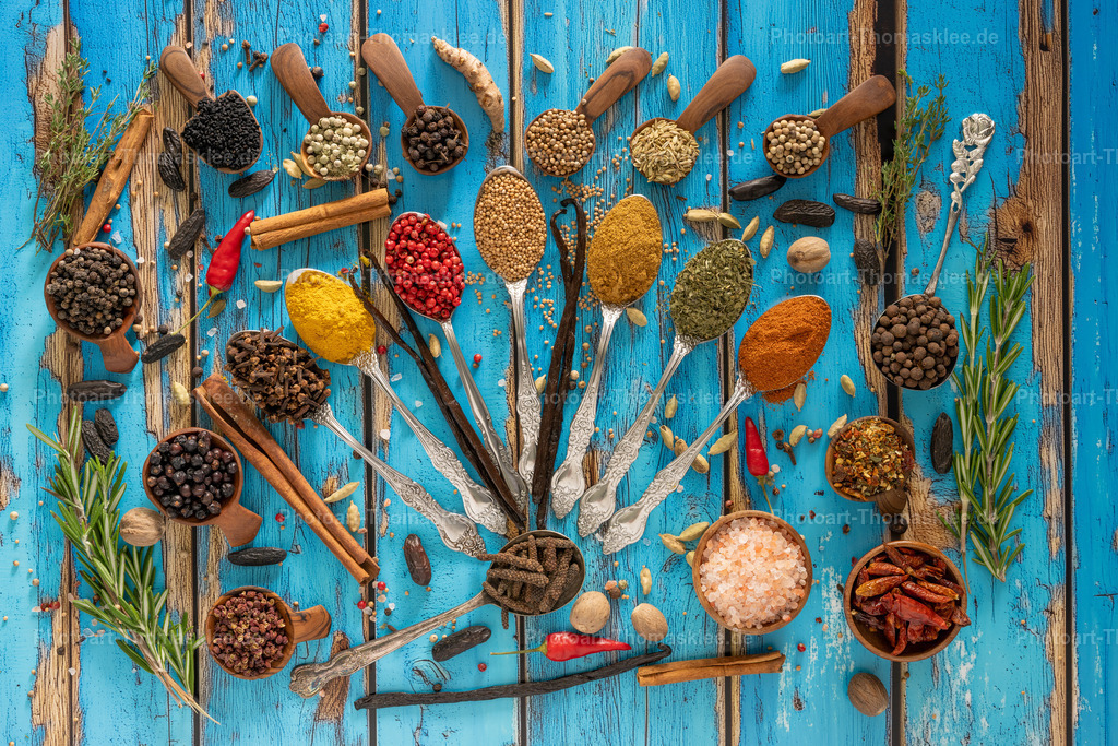 Gewürzvielfalt auf rustikalem Holz - Kulinarische Kunst I | Auf einem verwitterten blauen Holzuntergrund präsentiert sich eine kunstvolle Anordnung verschiedener Gewürze und Kräuter. Silberne Löffel und hölzerne Schaufeln tragen bunte Gewürze wie Kurkuma, Paprika, Pfeffer und getrocknete Chilis. Frische Rosmarinzweige rahmen die Komposition ein, während Zimt-Stangen und rosa Salz zusätzliche Farbakzente setzen. Diese harmonische Darstellung zeigt die Vielfalt der Küchenschätze und lädt zum Kochen ein. Die rustikale Optik verleiht dem Motiv einen authentischen Charme. - Realisiert mit Pictrs.com