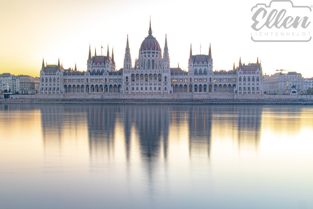 Golden Stillness | Bathed in the soft glow of morning light, the Hungarian Parliament reflects in perfect symmetry on the calm waters of the Danube. The city still slumbers, and for a brief moment, time seems to pause in golden silence. - Realisiert mit Pictrs.com