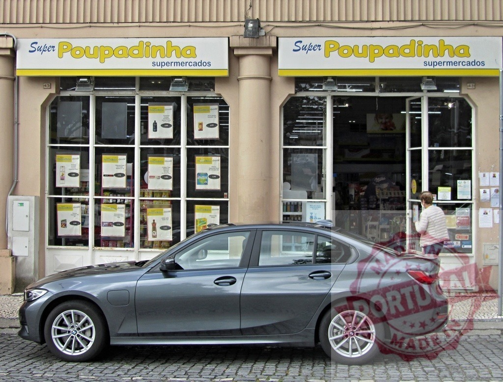 Super Poupadinha supermarket storefront with electric Car | Grey sedan electric car parked on a cobblestone street in front of a supermarket, with a person entering - Realized with Pictrs.com
