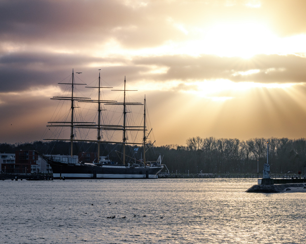 Die Passat im Sonnenlicht vor Warnemünde | Die Passat liegt vor der Küste von Warnemünde, während warme Sonnenstrahlen durch die Wolken brechen und das Wasser erhellen. Die dramatische Lichtstimmung verleiht der maritimen Szene eine besondere Tiefe. - Realisiert mit Pictrs.com