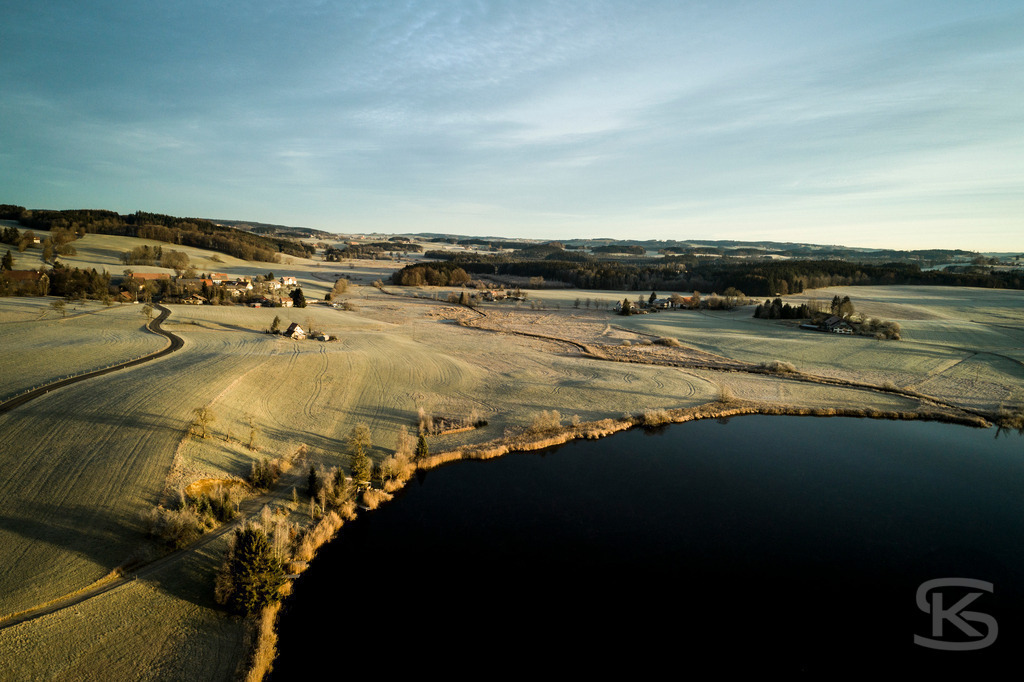 Winterliche Landschaft mit See bei Sonnenuntergang - Luftaufnahme Baden-Württemberg | Stimmungsvolle Drohnenaufnahme einer verschneiten Agrarlandschaft mit dunklem See im warmen Abendlicht. Idyllisches Winterpanorama mit gefrorenen Feldern, kleinem Dorf und sanften Hügeln in Süddeutschland. - Realisiert mit Pictrs.com