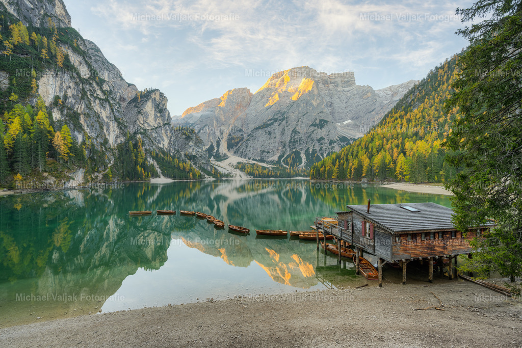 Pragser Wildsee in Südtirol an einem Herbstmorgen | Blick vom Ufer des Pragser Wildsees zu der Bootshütte und den aneinander gereihten Booten, die auf dem See treiben. Die Strahlen der frühen Morgensonne tauchen die Berggipfel in ein warmes Licht. - Realisiert mit Pictrs.com