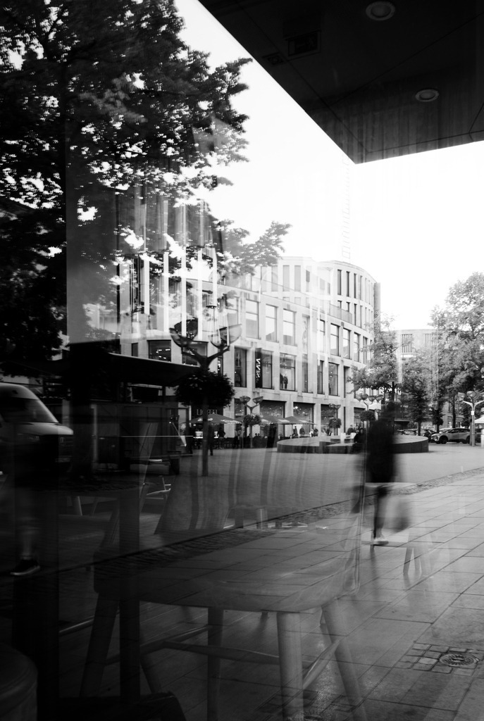 Zwischen Glas und Bewegung | Auf der Königstraße in Duisburg überlagern sich Architektur, Bäume und Cafémobiliar in einer Fensterscheibe. Eine vorbeigehende Person erscheint als flüchtige Bewegungsspur zwischen den klaren Linien der Gebäude.Die Schwarzweißaufnahme verbindet Spiegelungen und Durchblicke zu einer vielschichtigen Straßenszene, in der Innenraum und Außenwelt ineinanderfließen. - Realisiert mit Pictrs.com