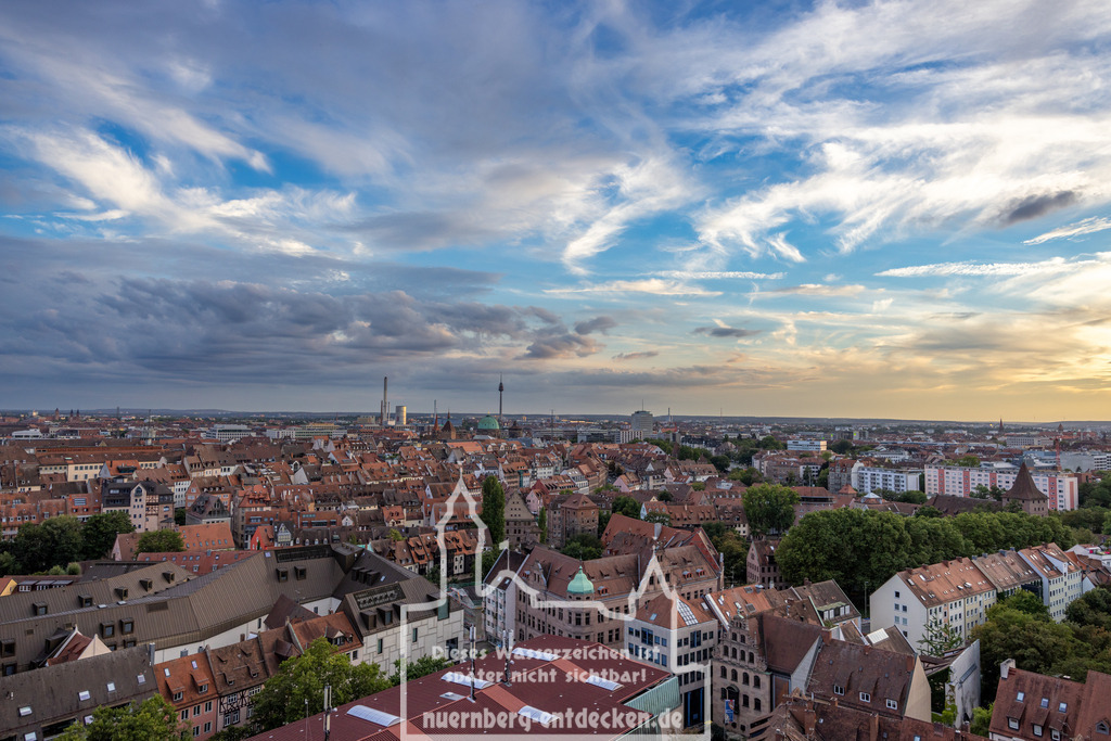 Nürnberger Stadtbild am Abend | Luftaufnahme der Nürnberger Altstadt von einem erhöhten Standpunkt. Die Dächer der historischen Gebäude erstrecken sich weit bis zum Horizont, wo Industrieanlagen und moderne Gebäude das Stadtbild ergänzen. Der Himmel zeigt eine beeindruckende Wolkenformation, während die untergehende Sonne das Panorama in warmes Licht taucht. - Realisiert mit Pictrs.com