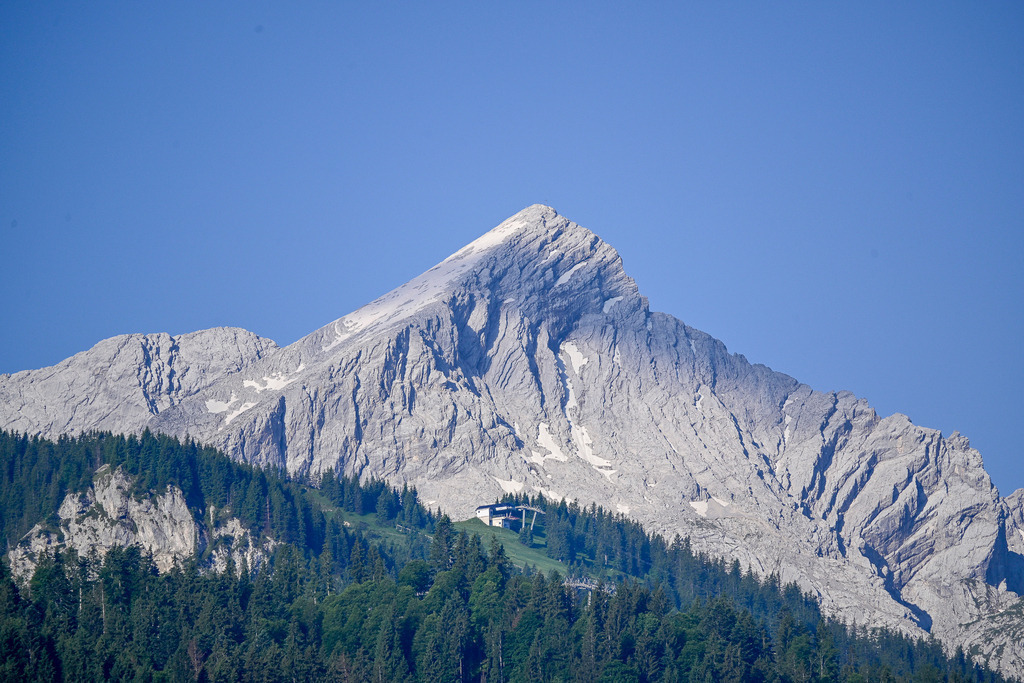 Steinerne Majestät im Sommerlicht: Die Alpspitze | Ein Anblick von zeitloser Erhabenheit und tiefer Ruhe: Stolz und mächtig ragt die charakteristische Pyramide der Alpspitze in den makellos blauen Sommerhimmel. Die schroffen, hellgrauen Felswände zeigen ihre faszinierende Struktur, in deren tiefen Rinnen noch letzte Schneereste aus dem Winter schimmern. Am Fuß des gigantischen Massivs schmiegt sich eine Bergstation wie ein winziger Außenposten an die saftig grünen Almwiesen und dichten Nadelwälder. Diese Aufnahme fängt die Ehrfurcht gebietende Kraft der Alpen ein und weckt eine unbändige Sehnsucht nach der Freiheit des Bergsommers. - Realisiert mit Pictrs.com