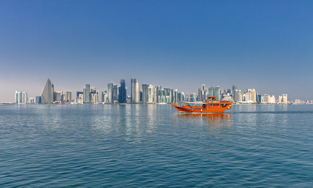 Skyline von Doha,Hauptstadt von Katar,Tradition trifft Moderne | Das Bild zeigt die moderne Skyline von Doha, der Hauptstadt von Katar, im Vordergrund mit einem traditionellen Holzboot (einer Dhau) im Persischen Golf.Die Aufnahme blickt von der See aus auf die markanten Wolkenkratzer der Finanz- und Geschäftsdistrikte entlang der Küste von Doha.Tradition trifft Moderne: Das orangefarbene Holzschiff symbolisiert das maritime Erbe des Landes, während die futuristische Hochhauskulisse den wirtschaftlichen Aufschwung der Region repräsentiert. - Realisiert mit Pictrs.com