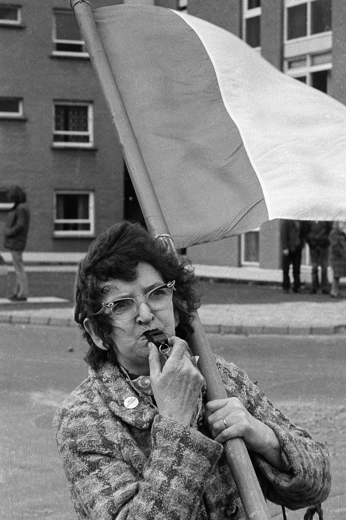 016-1973-009 | In der Bogside, dem katholischen Stadtteil von Derry/Londonderry: Szene am Rand einer Demonstration von Sinn Fein, dem politischen Arm der IRA. Die Trillerpfeife im Mund der Frau, die eine Fahne in den Farben Grün-Weiß-Orange als Zeichen der Unabhängigkeitsbestrebung von Großbritannien trägt, hat mehr als eine symbolische Bedeutung: Mit Mülleimerdeckeln und Trillerpfeifen warnten die Einwohnerinnen der katholischen Viertel Nordirlands die Kämpfer der IRA, wenn Soldaten der britischen Armee im Anmarsch waren. - Realisiert mit Pictrs.com
