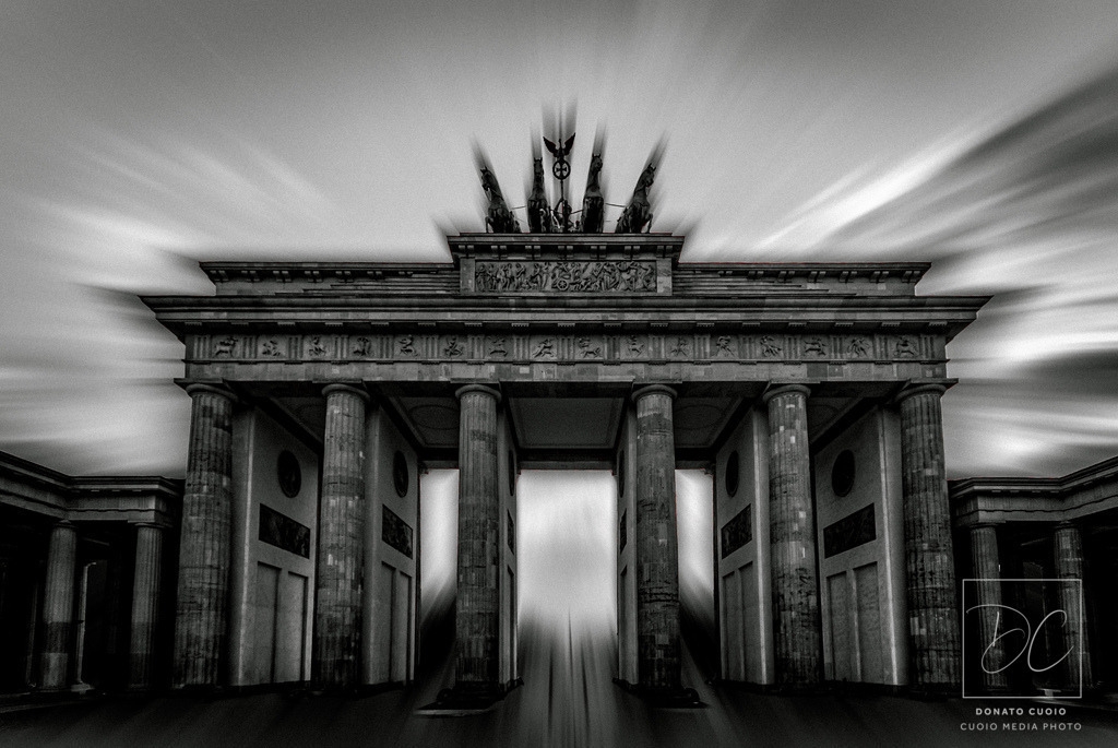 Das Brandenburger Tor – ein historisches Symbol neu interpretiert mit Licht und Dynamik | Das Bild zeigt das berühmte Brandenburger Tor in Berlin, jedoch mit einer künstlerischen, digitalen Bearbeitung. Es wurde in Schwarz-Weiß umgewandelt, was eine nostalgische oder dramatische Wirkung erzeugt. Durch den Einsatz eines Radialfilters, der von der Mitte des Bildes nach außen hin verläuft, entsteht eine dynamische, fast surreal wirkende Bildkomposition. Dies verleiht dem Foto eine gewisse Dramatik und zieht den Fokus auf das Zentrum des Bauwerks.Die Lichtstrahlen und Verzerrungseffekte verstärken den Eindruck von Bewegung, was eine fast mystische Atmosphäre erzeugt. Der hohe Kontrast zwischen den dunklen Säulen und dem hellen Himmel betont die architektonischen Details des Brandenburger Tors und verleiht dem Bild eine beeindruckende Tiefe. - Realisiert mit Pictrs.com