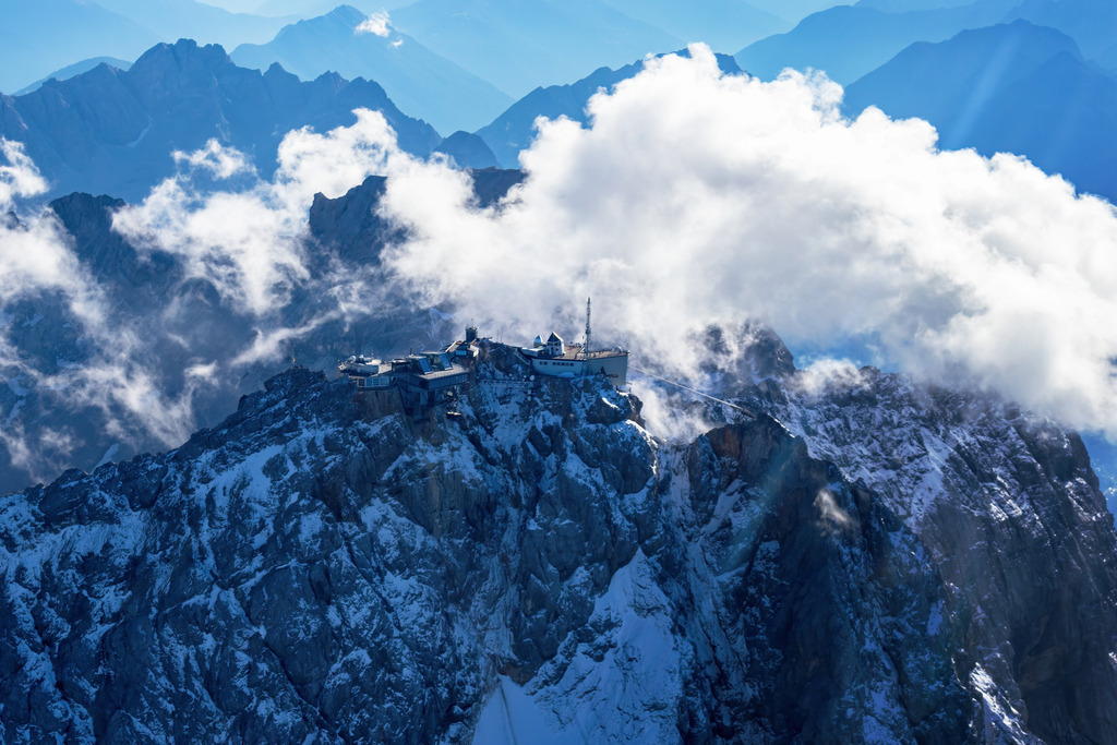 Wandbild - Majestätische Zugspitze | Ein atemberaubender Blick auf die Zugspitze, den höchsten Gipfel Deutschlands, der in eine dramatische Wolkendecke gehüllt ist. Die schneebedeckten Felsen und die schroffen Bergspitzen kontrastieren stark mit den weichen, weißen Wolken, die um die Gipfel schweben. Auf dem Gipfel befindet sich eine Bergstation mit mehreren Gebäuden, die die extremen Wetterbedingungen und die Höhe trotzen. Im Hintergrund erstrecken sich die imposanten Alpen, die in verschiedenen Blautönen zu sehen sind und eine majestätische Kulisse bilden.