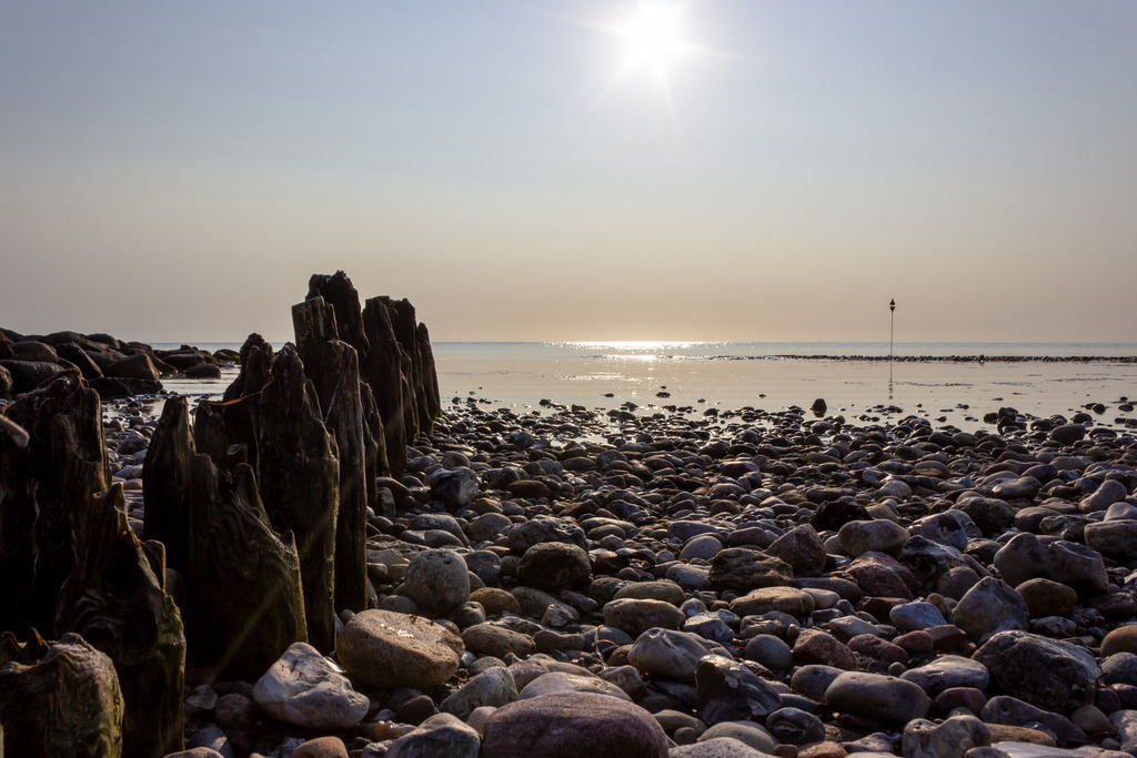 Wandbild: Sonnenschein am Naturstrand in Damp | Dieses Wandbild im Querformat zeigt schönen Sonnenschein am Naturstrand in Damp. Im Vordergrund sind Steine zu sehen. Die Sonnen spiegelt sich auf dem Meer.  - Realisiert mit Pictrs.com