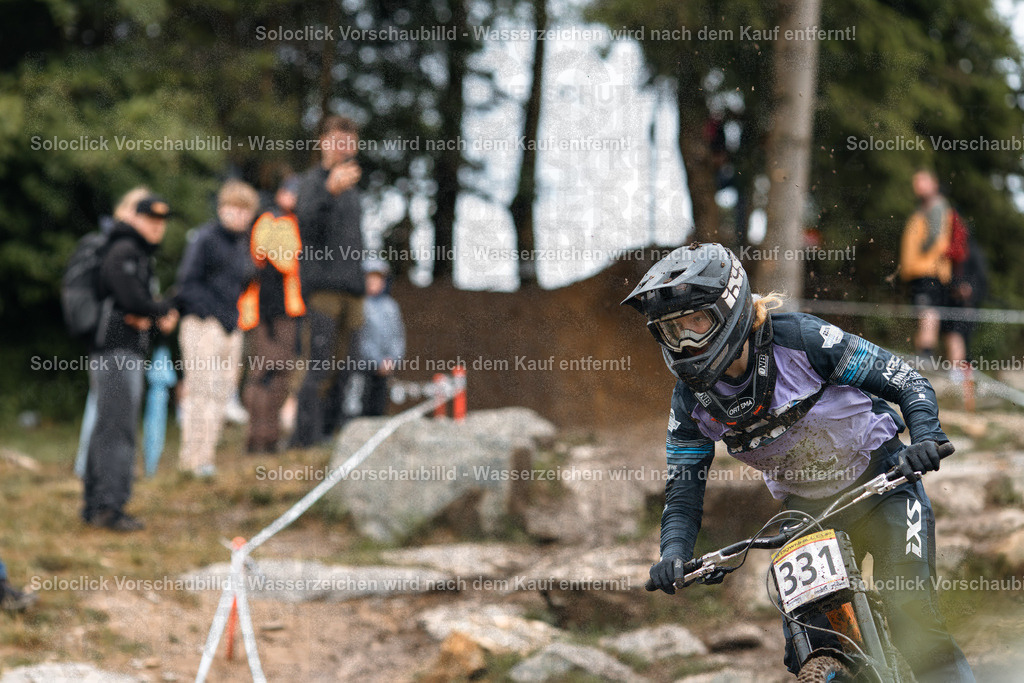 Mountainbike-Action im Wald | Das Bild zeigt einen Mountainbiker in voller Ausrüstung, der einen staubigen Pfad in einem Wald hinunterfährt. Er ist in einer dynamischen Pose, die Geschwindigkeit und Kontrolle vermittelt. Im Hintergrund sind Zuschauer zu sehen, die das Rennen beobachten. - Realisiert mit Pictrs.com