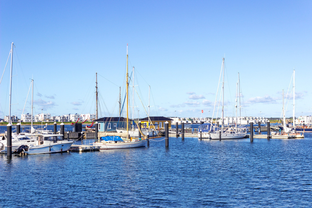 Wandbild: Yachthafen in Olpenitz | Dieses Wandbild im Querformat zeigt den Yachthafen in Olpenitz. Im Vordergrund ist das Wasser zu sehen. Im Hintergrund kann man die Ferienwohnungen in Olpenitz erahnen. Der blaue Himmel ist fast wolkenlos.  - Realisiert mit Pictrs.com