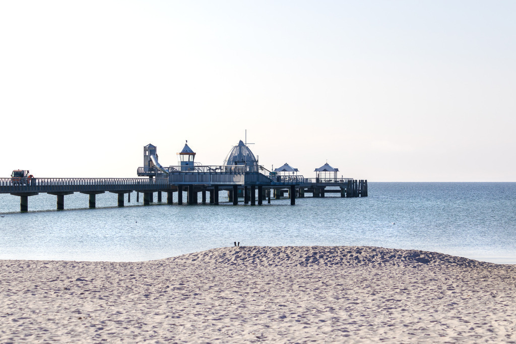 Wandbild: Weite und Wasser – Grömitz mit Seebrückenblick | Dieses Wandbild zeigt eine ruhige Szene vom Strand in Grömitz in den frühen Morgenstunden. Der Blick führt über den feinen, noch unberührten Sand hinaus auf das Meer, wo sich die Seebrücke mit ihren markanten Pavillons und dem kuppelartigen Bauwerk am Ende elegant ins Wasser erstreckt. Die Seebrücke fügt sich als architektonisches Element wirkungsvoll in die offene Strandkulisse ein. Das Licht ist weich und klar – typisch für den frühen Morgen – und taucht die Szene in eine friedliche, fast meditative Atmosphäre. Der wolkenlose Himmel verstärkt das Gefühl von Weite und Ruhe. Die Komposition wirkt strukturiert und zugleich weitläufig – ein Moment der Stille, bevor der Tag beginnt. Dieses Motiv eignet sich ideal als Wandbild für maritime Wohnkonzepte – ob als Leinwandbild, Acrylglasbild, Alu-Dibond FineArt Print oder als Akustikbild. Ein stilvoller Akzent für Wohnzimmer, Büro oder Ferienwohnung. - Realisiert mit Pictrs.com