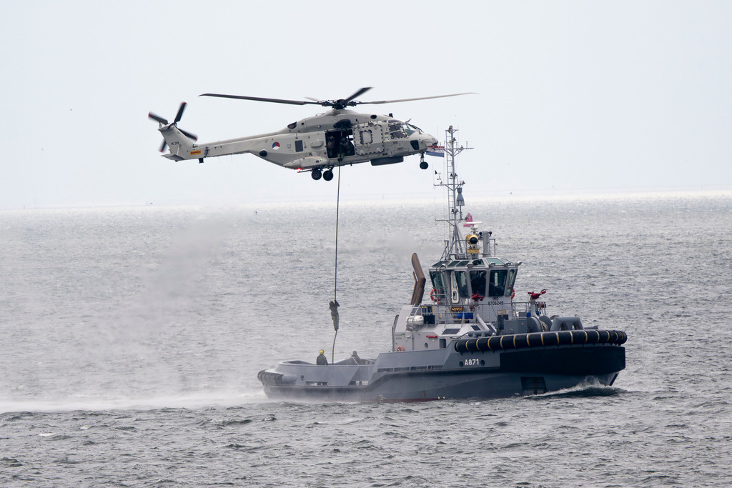 Winschmanöver auf See | Die Aufnahme dokumentiert eine dynamische Rettungsübung oder Abseilaktion auf offener See. Ein militärischer Hubschrauber (NH90), Typ Sealion der niederländische Marineflieger schwebt stabil über einem Schlepper der Küstenwache (A871), während sich eine Person an einem Seil zum Deck hinunterlässt. Das aufgewühlte Wasser und die Gischt unterstreichen die Präzision des Manövers. - Realisiert mit Pictrs.com