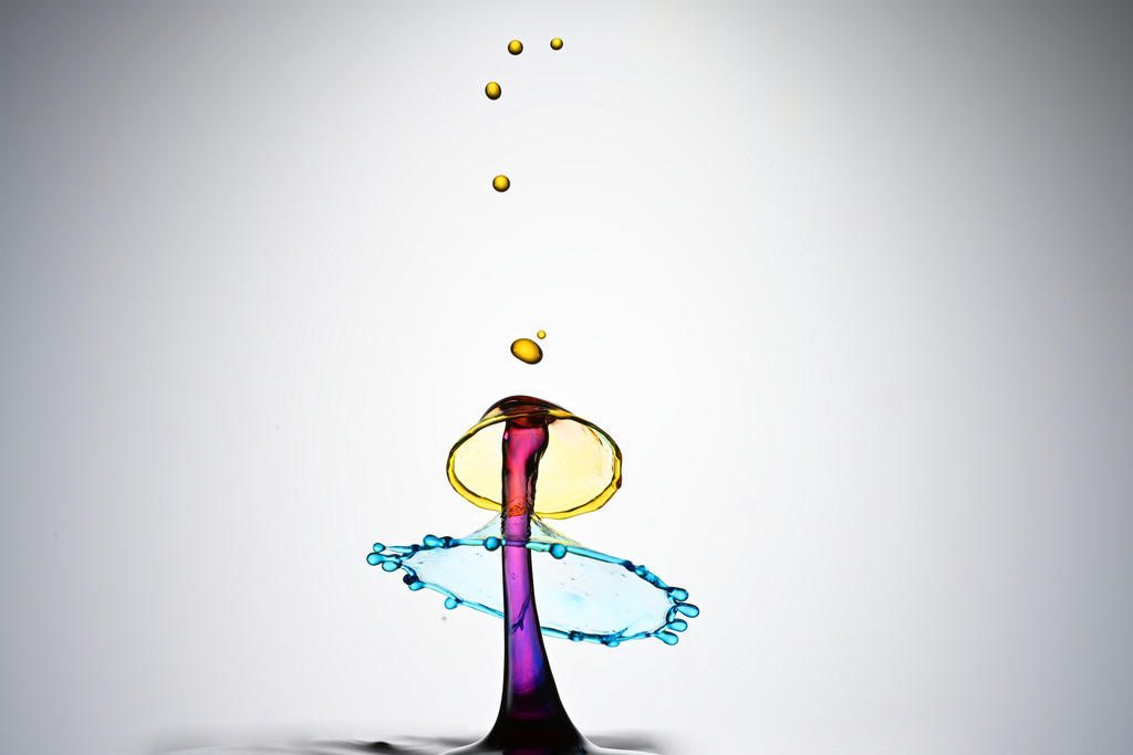 Fantastisches Wassertropfenkollision | Erleben Sie die atemberaubende Schönheit der High-Speed-Fotografie mit dieser faszinierenden Wassertropfen-Kollision. Das Bild fängt den perfekten Augenblick ein, in dem zwei Tropfen aufeinandertreffen und eine pilzförmige Flüssigkeitssäule bilden, umgeben von einem leuchtend blauen Spritzkranz. Die intensiv gefärbten Flüssigkeiten – leuchtendes Gelb (oder Gold) und brillantes Blau – erzeugen einen dramatischen Kontrast vor einem neutralen Hintergrund. Ideal für Projekte, die Dynamik, Wissenschaft, Physik, Makrofotografie oder abstrakte Schönheit darstellen sollen. Ein beeindruckendes Beispiel für Tropfenkunst und Flüssigkeitsfotografie.