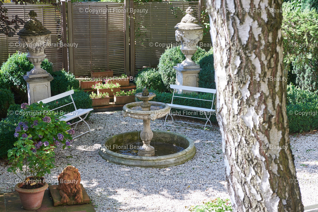 Mediterraner Stadtgarten | Brunnen in Stadtgarten mit Kies, weißen Gartenbänken und antiken Amphoren. - Realized with Pictrs.com