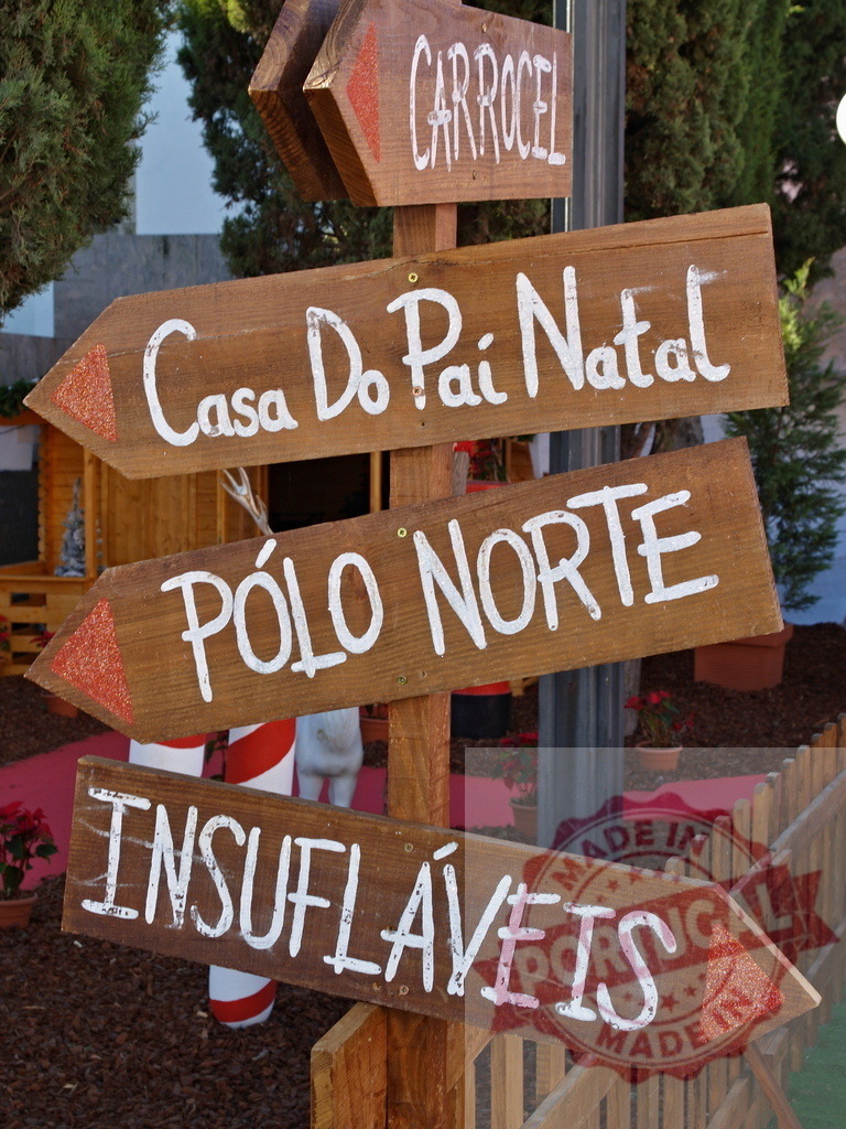 Wooden signpost pointing to north pole, santa claus house, carousel and inflatable games | Wooden direction signs in portuguese language pointing to north pole, santa claus house, carousel and inflatable games in a christmas theme park - Realisiert mit Pictrs.com
