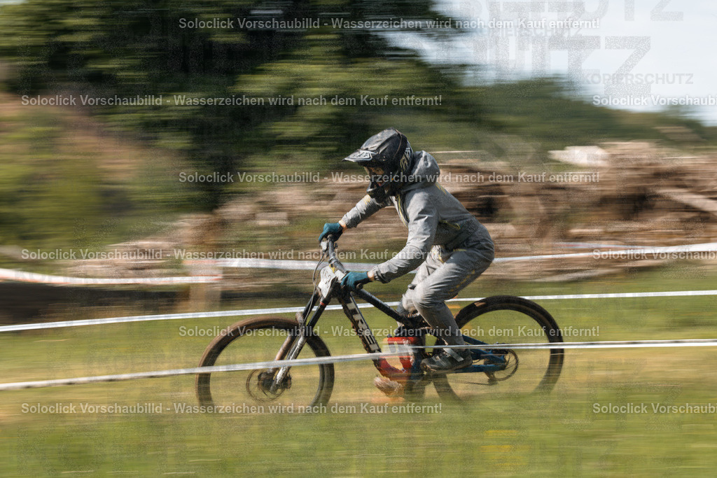 Mountainbike-Action | Ein Mountainbiker in voller Ausrüstung rast auf einem Feld durch, wobei die Bewegung durch eine lange Belichtungszeit dargestellt wird. Der Hintergrund besteht aus Bäumen und einem Hügel. - Realisiert mit Pictrs.com