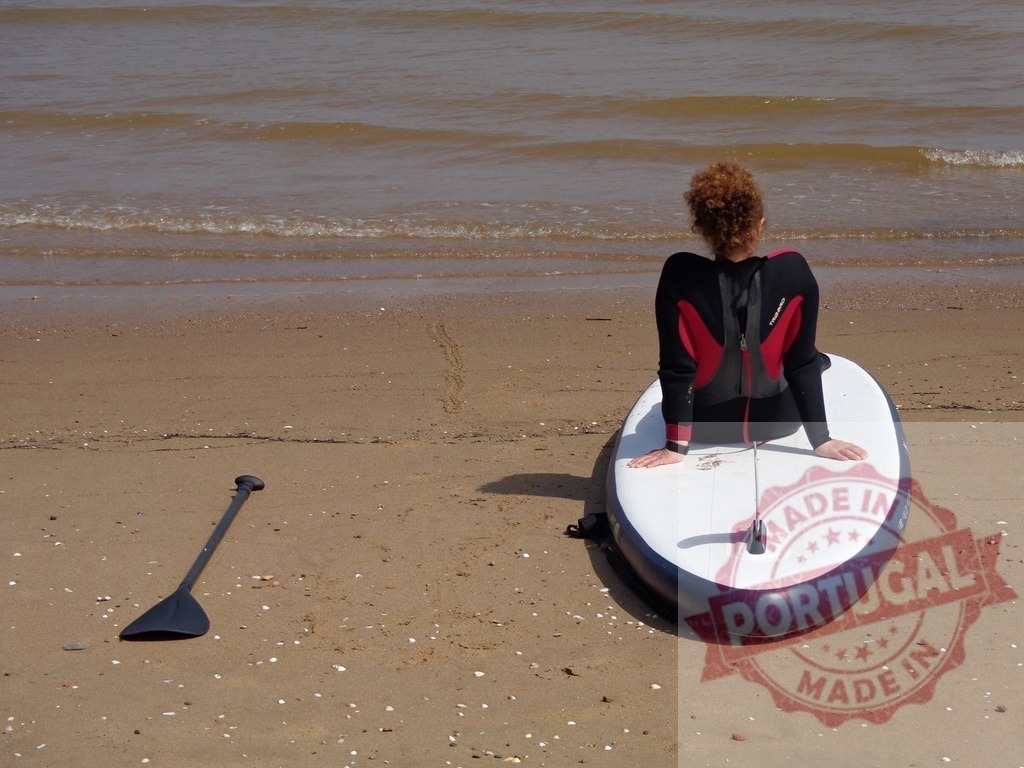 Woman in wetsuit sitting on paddleboard on beach | Woman in wetsuit sitting on paddleboard on beach looking at the ocean - Realisiert mit Pictrs.com