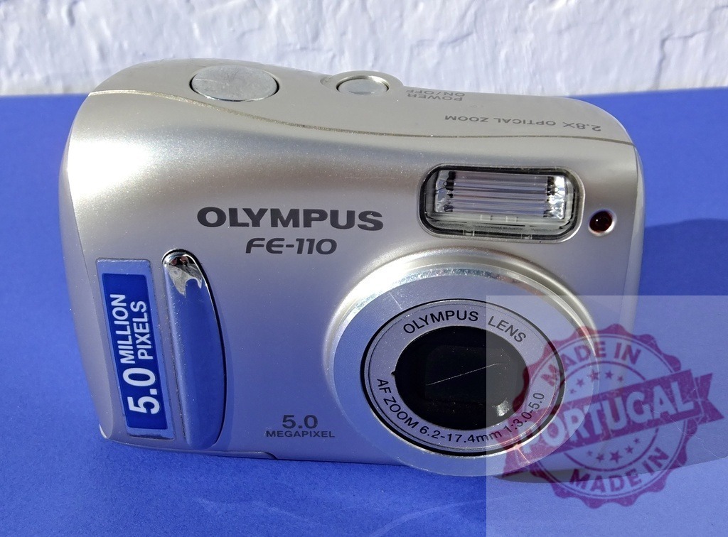 Olympus FE-110 silver compact digital camera | Olympus FE-110, a silver compact digital camera, sitting on a blue background - Realized with Pictrs.com