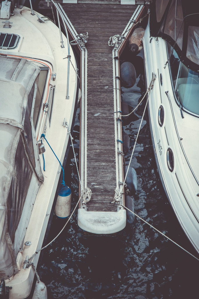 Between the Boats | Ein schmaler Steg liegt zwischen zwei Booten und wird aus der erhöhten Perspektive zum Mittelpunkt einer klar gegliederten maritimen Komposition.Die symmetrische Anordnung, verwittertes Holz, Taue und dunkles Wasser erzeugen eine ruhige, leicht nostalgische Atmosphäre. Die zurückhaltenden Blau-, Grau- und Cremetöne verleihen dem Motiv einen modernen Moody-Look und betonen zugleich die grafische Struktur der Szene.Ein maritimes Fine-Art-Motiv zwischen Geometrie, Ruhe und Hafenleben – ideal für moderne, minimalistische Wohnräume. - Realisiert mit Pictrs.com