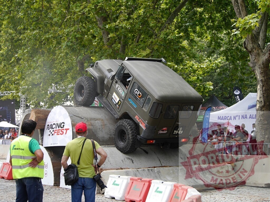 Four-wheel drive vehicle climbing concrete ramp during off-road competition | Modified jeep climbing a steep concrete ramp during an off-road racing competition, watched by spectators and supervised by security personnel - Realized with Pictrs.com
