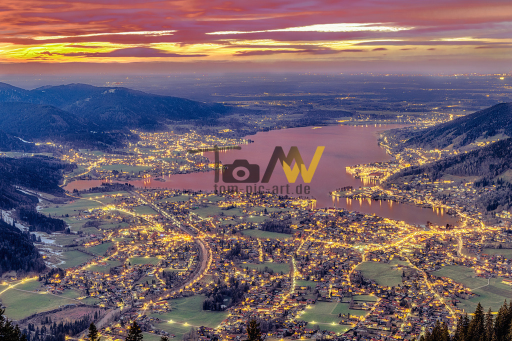 Tegernsee bei Nacht von oben | Nachtaufnahme des Tegernsee , von oben im Licht der untergehenden Sonne. Straßenlichter.Die Abbildung zeigt eine Luftaufnahme des Tegernsees bei Dämmerung oder Sonnenuntergang, umgeben von den beleuchteten Ortschaften im Tegernseer Tal. Der Tegernsee liegt etwa 50 Kilometer südlich von München in den Bayerischen Voralpen und ist ein beliebtes Ausflugs- und Urlaubsziel. Die gleichnamige Stadt Tegernsee liegt am Ostufer des Sees. Der See gilt als eines der saubersten Gewässer Europas, was auf die in den 1960er Jahren installierte Ringkanalisation zurückzuführen ist. Ansehnliche Berge wie der 1722 Meter hohe Wallberg umrahmen den See.  - Realisiert mit Pictrs.com