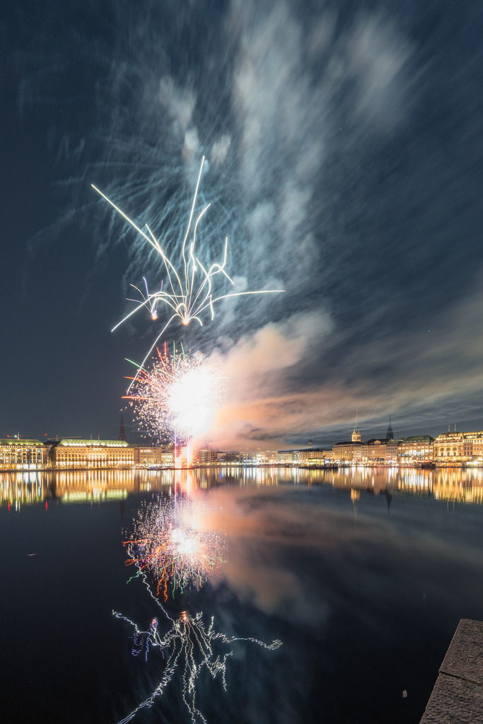 Feuerwerk im Hamburger Nachthimmel | Helle Feuerwerksbahnen durchbrechen die dramatischen Wolken über der Alster, während ihre Lichtspuren auf der Wasseroberfläche reflektiert werden. - Realisiert mit Pictrs.com