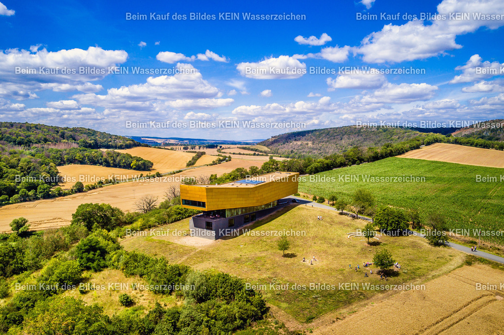 Besucherzentrum Arche Nebra - die Himmelsscheibe-0012 | Dieses Luftbild zeigt die Arche Nebra im Burgenlandkreis in Sachsen-Anhalt an einem sonnigen Sommertag. Das moderne Besucherzentrum liegt auf dem Mittelberg bei Nebra und ist eng mit einem der bedeutendsten archäologischen Funde Europas verbunden – der Himmelsscheibe von Nebra. Aus der Vogelperspektive werden die markante Architektur des Museums und die weitläufige Landschaft der Region besonders eindrucksvoll sichtbar.Die Arche Nebra wurde als Informations- und Erlebniszentrum rund um die Himmelsscheibe errichtet, die als älteste konkrete Darstellung des Sternenhimmels gilt. Das außergewöhnliche Gebäude fügt sich harmonisch in die sanften Hügel und Felder der Saale-Unstrut-Region ein und bildet einen modernen Kontrast zur historischen Bedeutung des Fundortes.Besonders beeindruckend ist die weite Landschaft des südlichen Sachsen-Anhalts, die sich rund um das Museum erstreckt. Felder, Wiesen und kleine Waldflächen prägen das Bild und vermitteln einen Eindruck von der ursprünglichen Kulturlandschaft, in der die Himmelsscheibe vor rund 3.600 Jahren verborgen wurde.Dieses Luftbild zeigt die Arche Nebra als eines der bedeutendsten archäologischen Besucherzentren Deutschlands und vermittelt einen faszinierenden Blick auf Geschichte, Wissenschaft und die reizvolle Landschaft der Saale-Unstrut-Region. - Realisiert mit Pictrs.com
