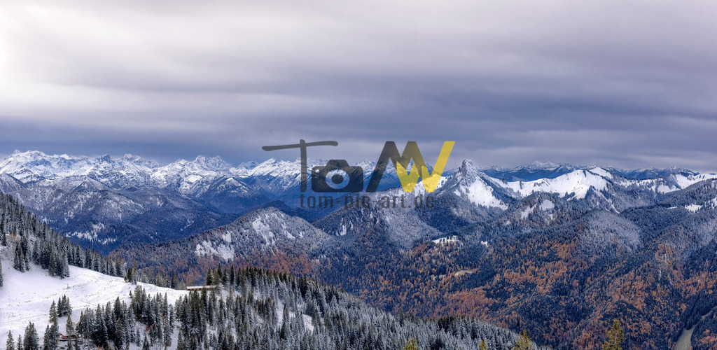 Winterliches Berg-Panorama--Wallberg--Horizont---Tegernsee  | Panorama über die bayrische winterliche Alpenkulisse am Tegernsee. Berge soweit das Auge reicht. Wunderschöne FernsichtDas Bild zeigt eine Winterlandschaft in den Bayerischen Alpen. Die Aufnahme ist ein Panoramafoto, das eine weite Bergkette mit schneebedeckten Gipfeln und Hängen zeigt.Im Vordergrund sind bewaldete Hänge mit Nadelbäumen zu sehen, die teils verschneit sind.Der Himmel ist bedeckt mit grauen und blauen Wolken. - Realisiert mit Pictrs.com