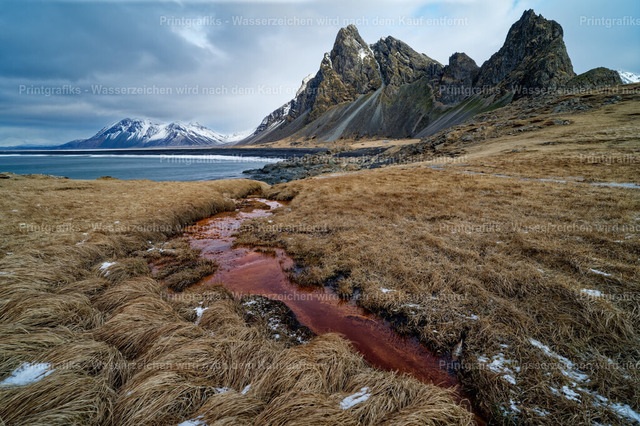 Raue Landschaft bei Stokksnes in Island | Raue Islandlandschaft am Vestrahorn mit trockenem Gras, dunklen Bergspitzen und einem rötlich gefärbten Bachlauf im Vordergrund. Die kühle Stimmung, die dramatischen Felsen und die offene Weite zeigen die ursprüngliche Schönheit der isländischen Küste. - Realisiert mit Pictrs.com