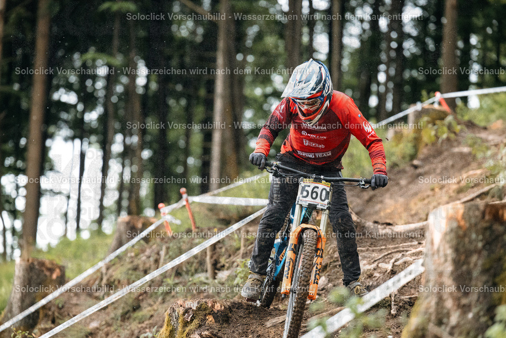 Mountainbiker im Rennen | Ein Mountainbiker rast einen steilen, schlammigen Weg hinunter, umgeben von dichtem, grünen Wald. Der Fahrer trägt einen roten Helm und ein rotes Trikot mit Sponsorenlogos. Er fährt ein Mountainbike mit roten und schwarzen Rahmen. Der Weg ist mit roten und weißen Bändern markiert, die den Kurs abgrenzen. Der Hintergrund besteht aus hohen, grünen Bäumen, die den Weg säumen. Es regnet leicht, was den Weg rutschig und schlammig macht. - Realisiert mit Pictrs.com