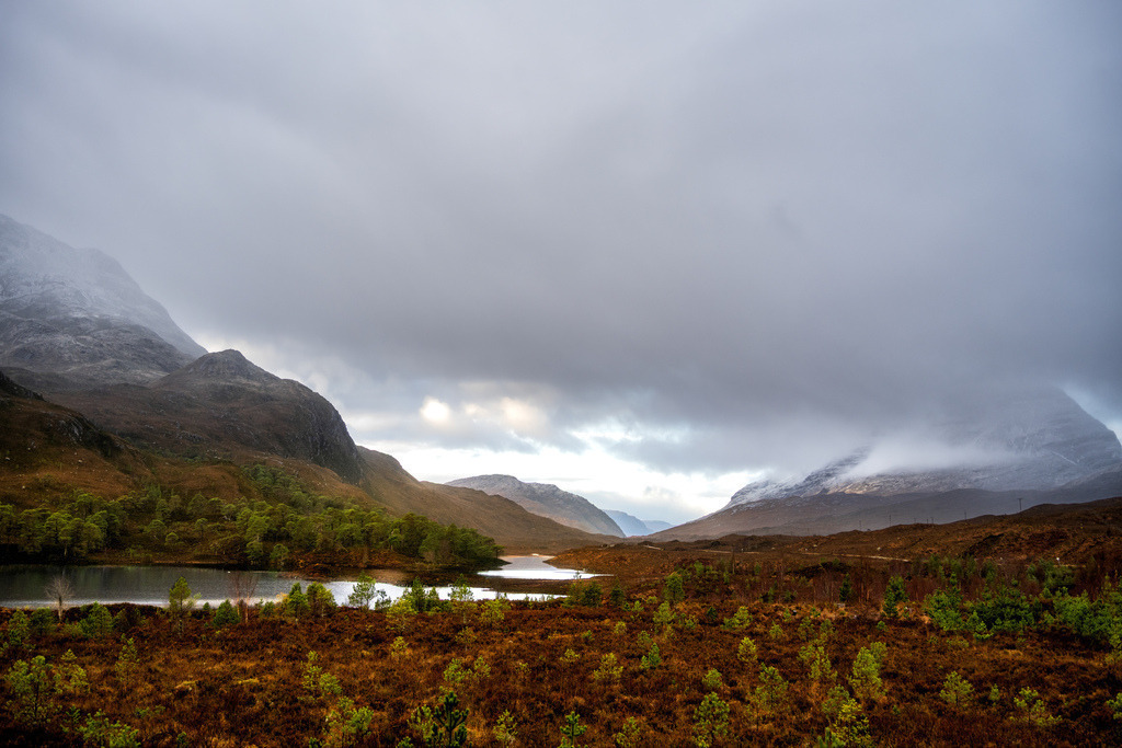 Loch Clair: Schottische Berglandschaft | A braid, roch mountain-landskaip unner a heavy, clood-happit lift. In the foregrund raxes a broun-reid muirland wi a wheen o' sma, bricht-green fir trees growin here an thair. Tae the left, a wee, derk loch sits at the fit o' a widit brae. In the hintgrund, muckle, snaw-tappit bens rise up, whiles biddin lown in the thick, gray cloods. A glint o' licht brakks throu the cloods ower the glen in the middle, giein a bit o' brichtness tae the dowie, dreich air.Eine weite Landschaftsansicht von Loch Clair in den schottischen Highlands zeigt einen ruhigen See, umgeben von sanften Hügeln und Bergen. Die herbstliche Moorlandschaft im Vordergrund präsentiert erdige Brauntöne und grüne Nadelbäume, während im Hintergrund nebelverhangene Gipfel unter einem bewölkten Himmel liegen. Die Komposition betont die Weite und raue Schönheit der Natur. - Realisiert mit Pictrs.com