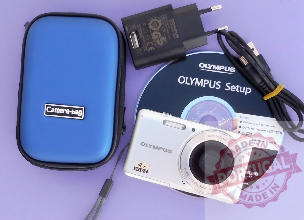 Olympus digital camera kit with accessories on purple background | Olympus digital camera and essential accessories including a blue camera case, charger, and setup CD - Realized with Pictrs.com