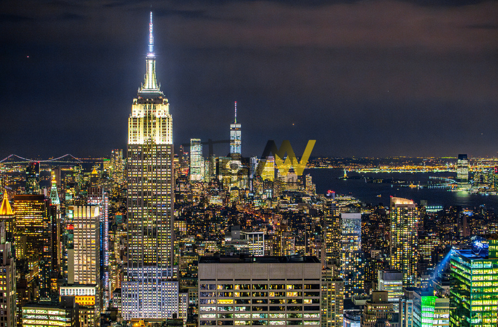 Skyline von New York-------Empire State Building bei Nacht | Das Bild zeigt eine atemberaubende Nachtaufnahme der New Yorker Skyline mit dem prominent beleuchteten Empire State Building im Mittelpunkt. Die Aufnahme wurde vom Top of the Rock im Rockefeller Center aus gemacht, da man von dort aus den besten Blick auf das Empire State Building selbst hat. Details zur SzeneStandort der Aufnahme: Die Perspektive lässt darauf schließen, dass das Foto von der Aussichtsplattform des Rockefeller Centers, bekannt als "Top of the Rock", aufgenommen wurde, da das Empire State Building vollständig sichtbar ist.Beleuchtung: Das Empire State Building ist nachts beleuchtet und dient als zentraler Blickfang der Szene. Die Lichter des Turms werden typischerweise bis 2 Uhr morgens ausgeschaltet.Atmosphäre: Die Szene fängt die lebendige und helle Atmosphäre der "Stadt, die niemals schläft" ein, mit unzähligen Lichtern der umliegenden Wolkenkratzer und der fernen Skyline von Lower Manhattan im Hintergrund.  - Realisiert mit Pictrs.com