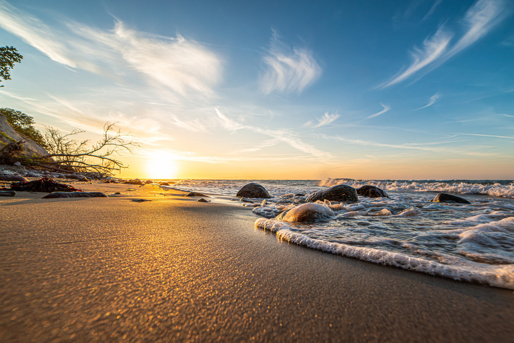 Sonnenuntergang am Strand von Stohl | Im Vordergrund des Bildes erstreckt sich das ruhige Meer mit sanften Wellen, die sanft gegen den Strand branden. Entlang des Strandes befinden sich vereinzelt Steine, die vom Wasser geformt wurden. Sie bilden interessante Kontraste zur Weichheit des Sandes und verleihen der Küstenlinie eine markante Struktur.